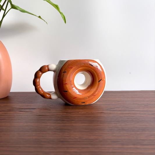 Ceramic donut-shaped mug on a wooden surface with a white wall and plant in the background