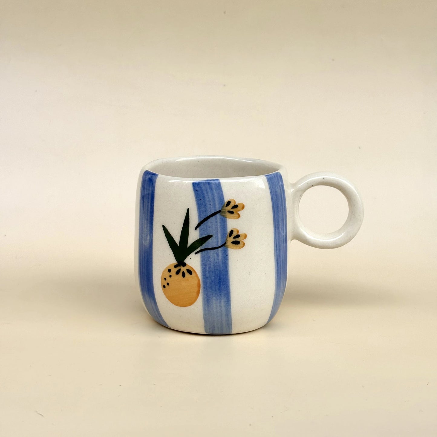 A small white ceramic mug with blue vertical stripes and a hand-painted pineapple design on the front, set against a plain beige background.