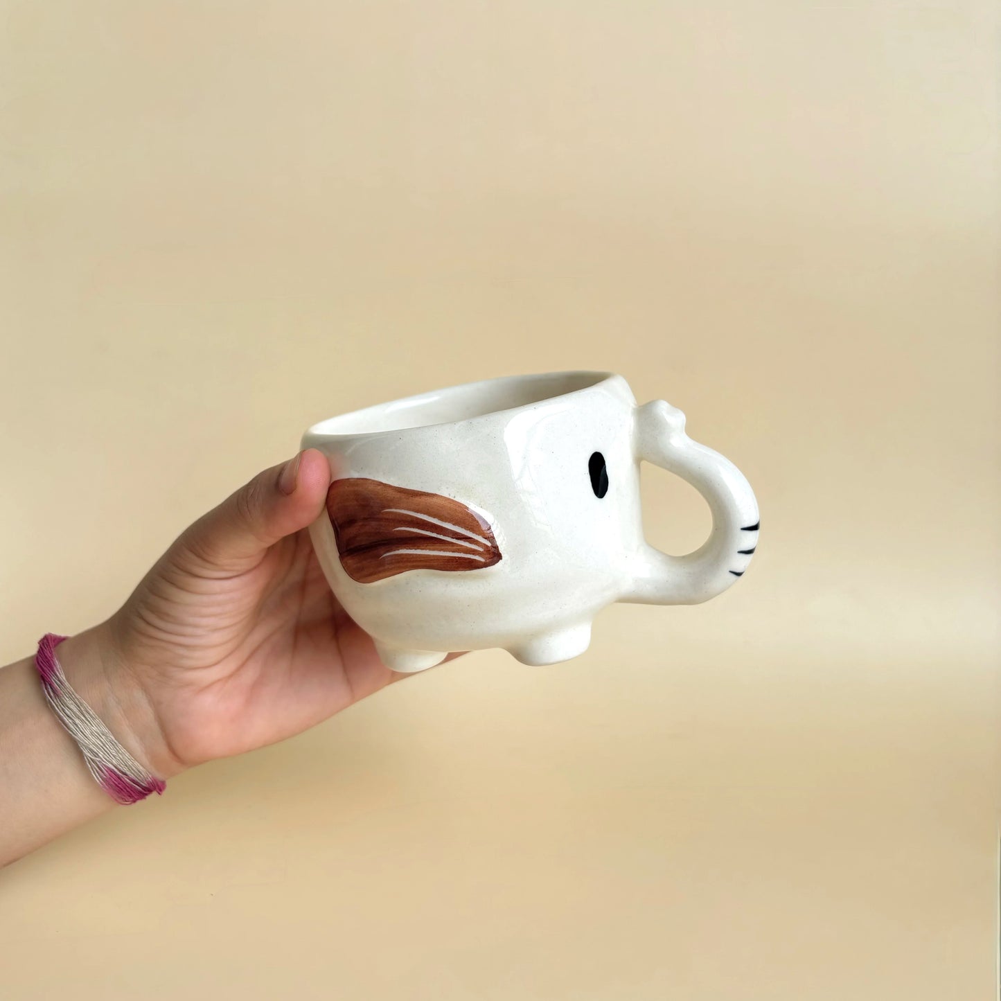 Hand holding a white ceramic mug shaped like an elephant with brown trunk on a beige background