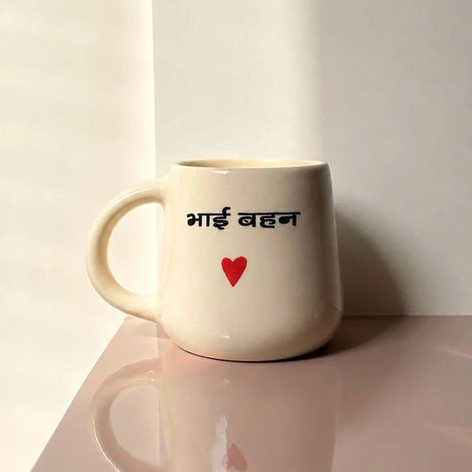 A cream-colored ceramic mug with the Hindi text 'Bhai Bahen' in black and a small red heart printed on the side, set on a reflective surface against a neutral background.