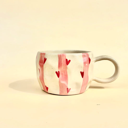 A handmade ceramic mug with a textured off-white body, decorated with vertical pink stripes and small red hearts, set against a plain cream background.