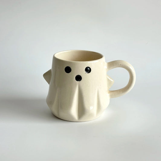 White ceramic mug shaped like a ghost with black eyes and mouth on a light gray background