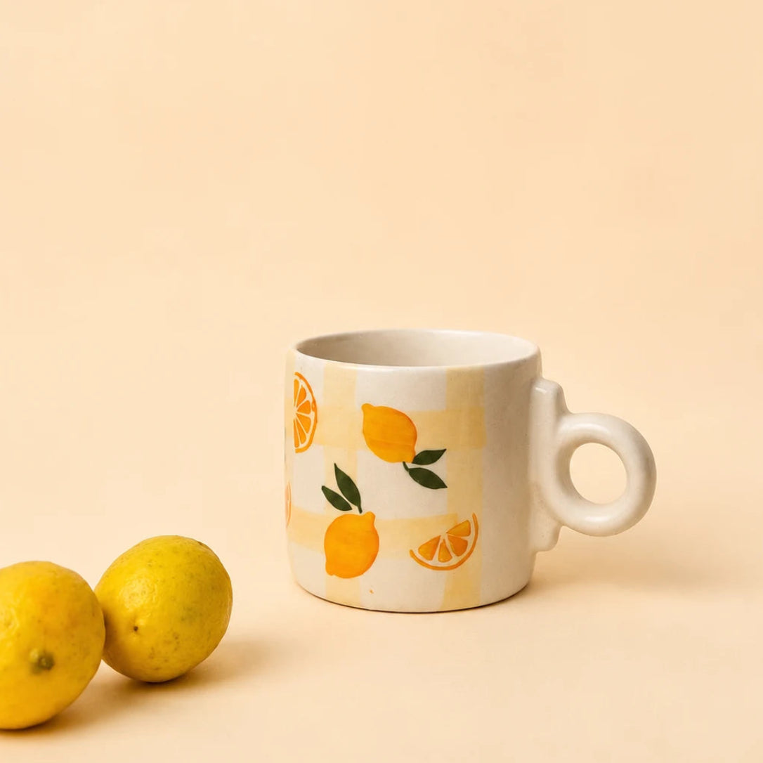 A white ceramic mug with a handle, decorated with a pattern of lemons and orange slices, set against a warm beige background with two lemons on the left.
