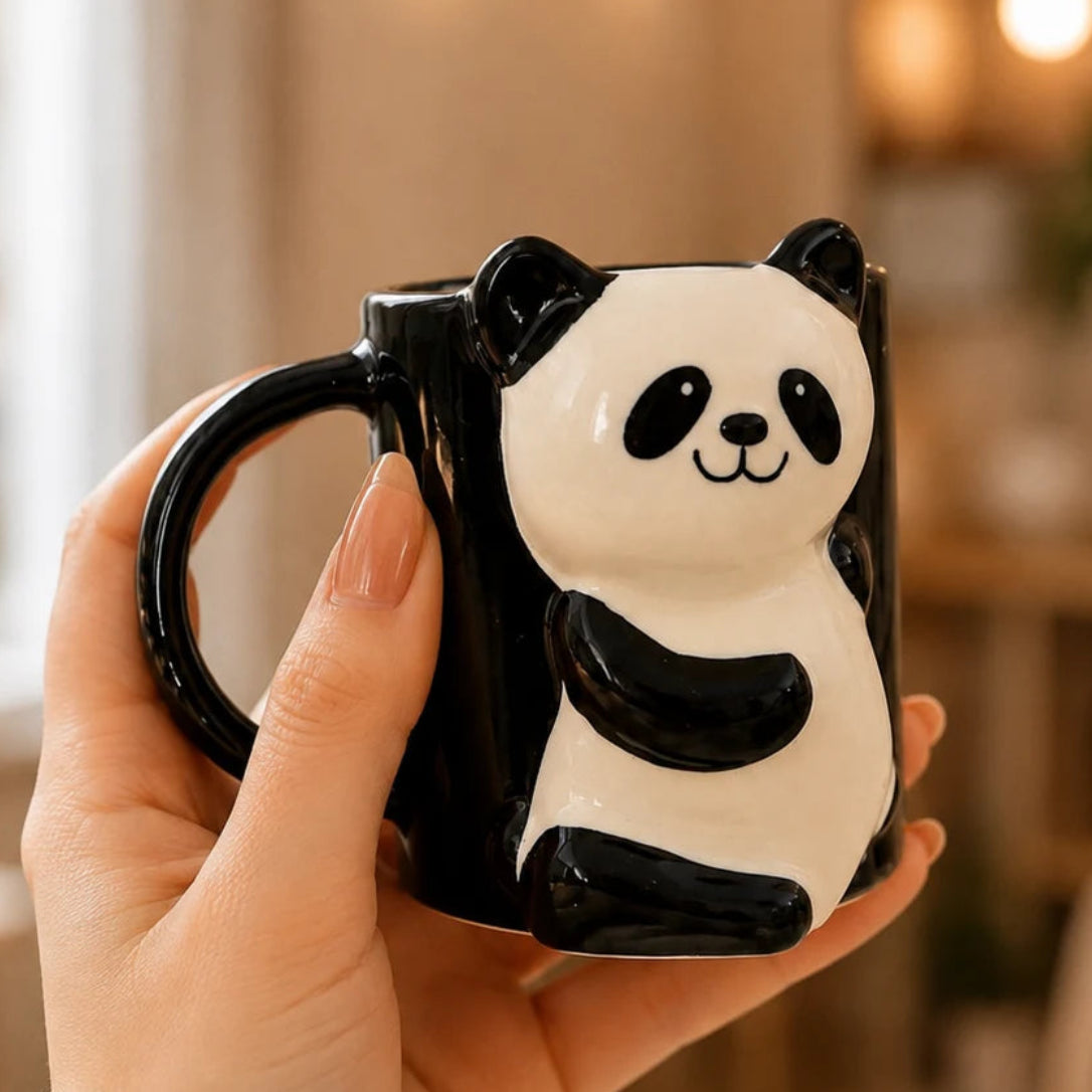 A hand holding a black and white ceramic mug shaped like a smiling panda, set against a softly blurred warm indoor background.