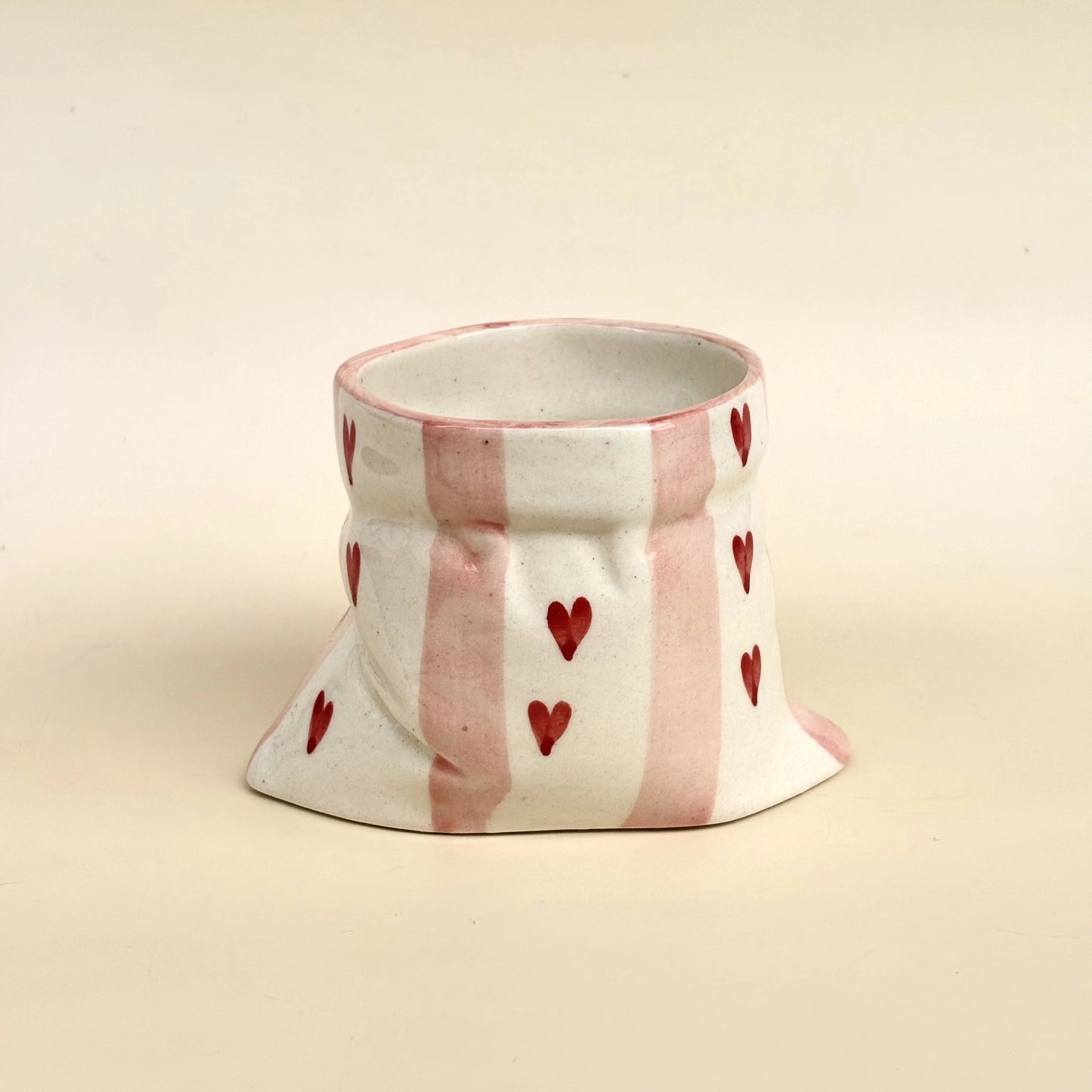 A handmade ceramic cup with a flared base, decorated with vertical pink stripes and small red hearts on a cream-colored glaze, set against a solid cream background.
