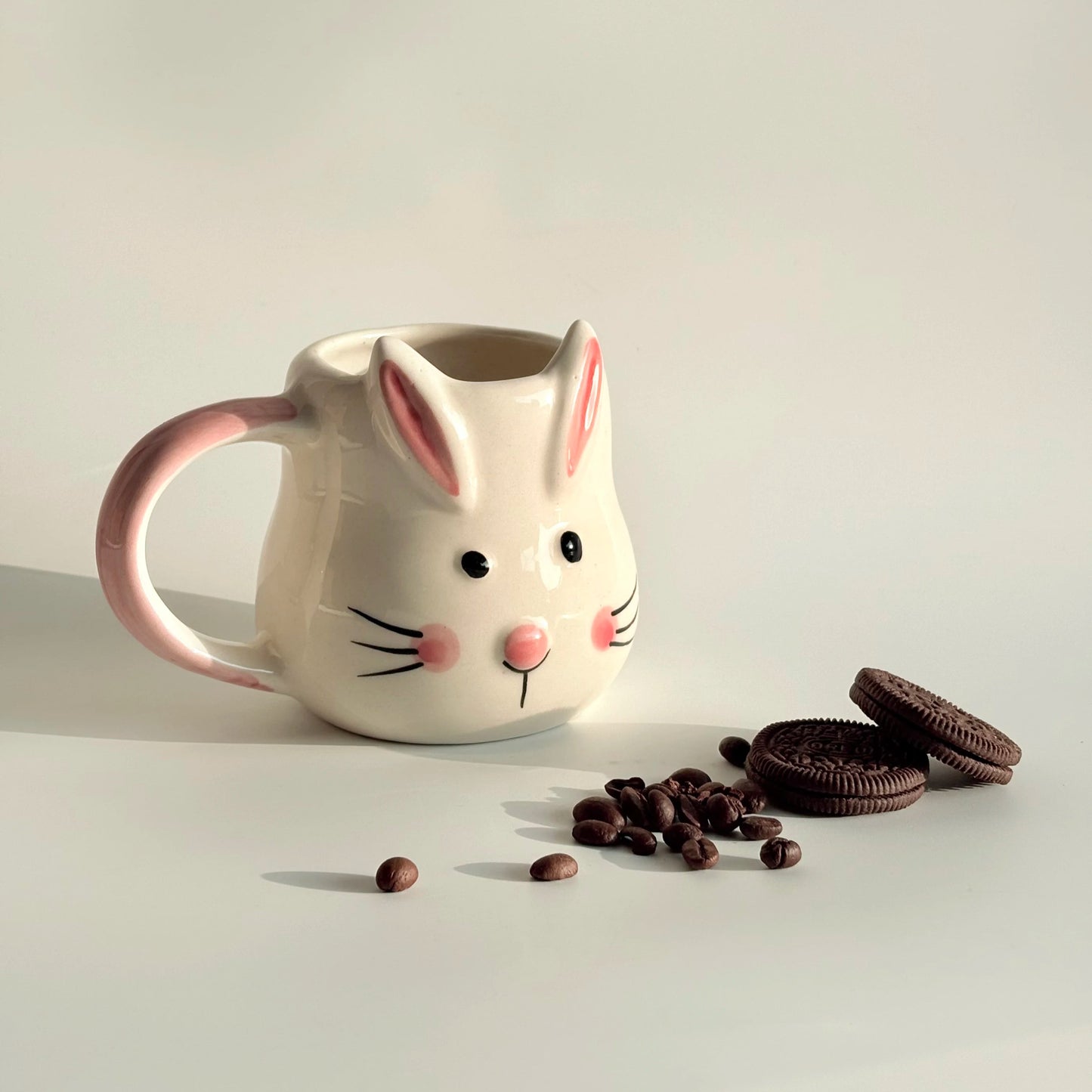Ceramic mug shaped like a rabbit with cookies and coffee beans on a light background