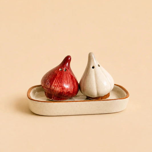 Two ceramic garlic-shaped salt and pepper shakers, one red and one off-white, sitting on a matching beige ceramic tray against a solid beige background.