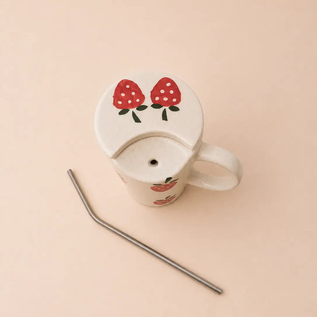 A white ceramic mug with a lid decorated with red strawberries, shown with a metal straw on a solid pale peach background.
