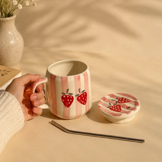 A ceramic mug with pink and white vertical stripes and red strawberry illustrations, held by a hand. The matching lid, a metal straw, and a vase of baby's breath are arranged on a beige surface with soft leaf shadows.