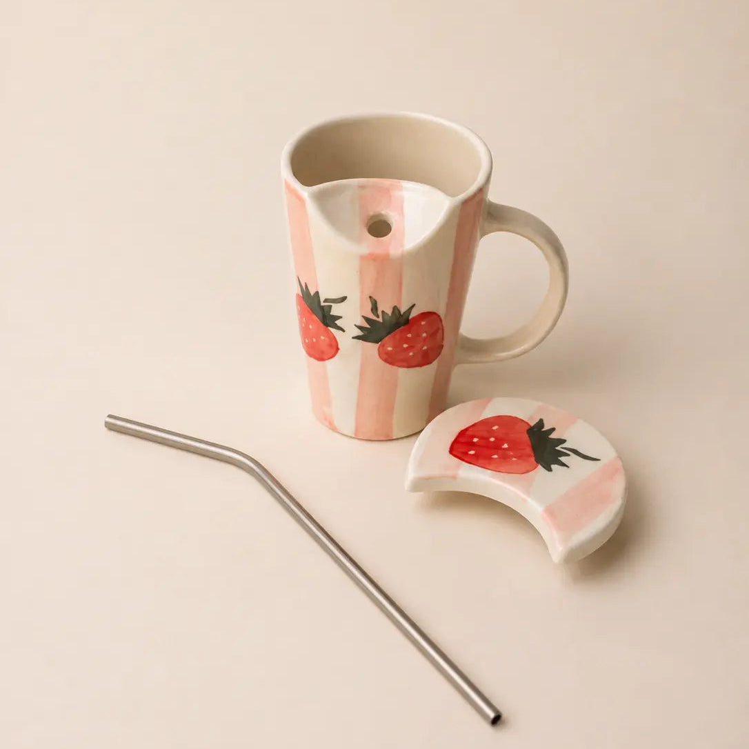 A ceramic mug with a handle, decorated with pink and white vertical stripes and red strawberries, shown with a matching lid and a metal straw on a light beige background.