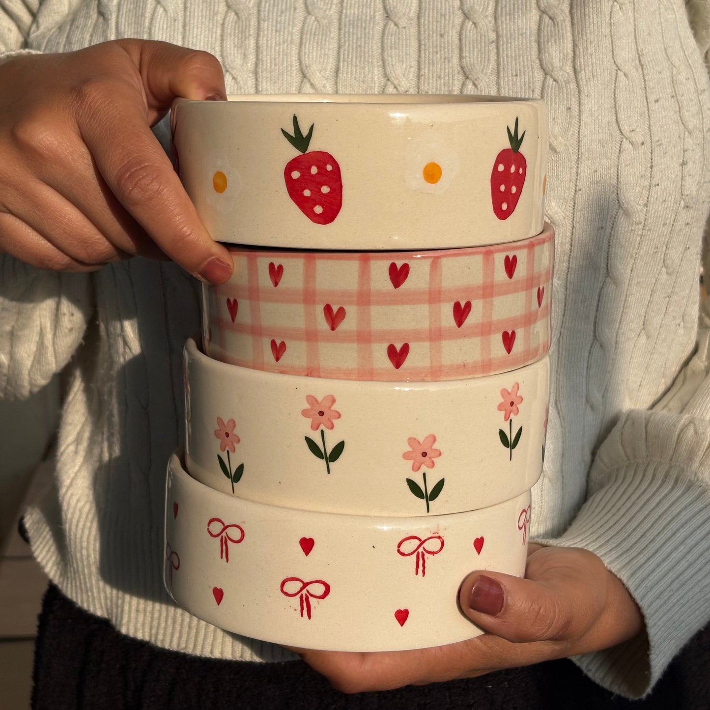 A person wearing a cream cable-knit sweater holds a stack of four round ceramic bowls decorated with cute patterns. The bowls feature strawberry, heart, flower, and bow motifs in red, pink, and green on a warm cream background.
