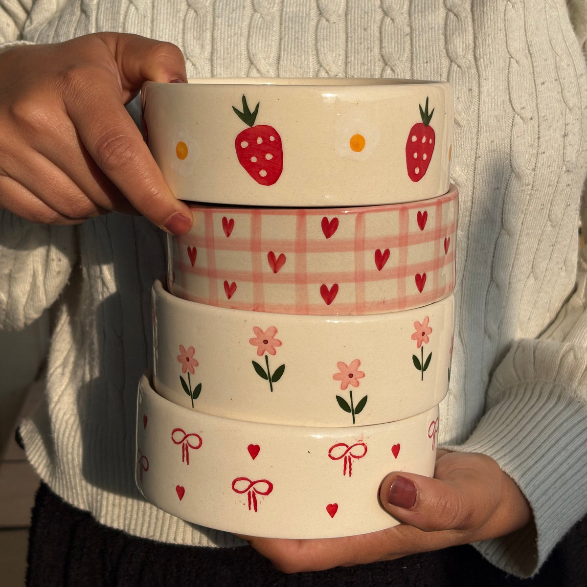 A person wearing a cream cable-knit sweater holds a stack of four round ceramic bowls decorated with cute patterns. The bowls feature strawberry, heart, flower, and bow motifs in red, pink, and green on a warm cream background.