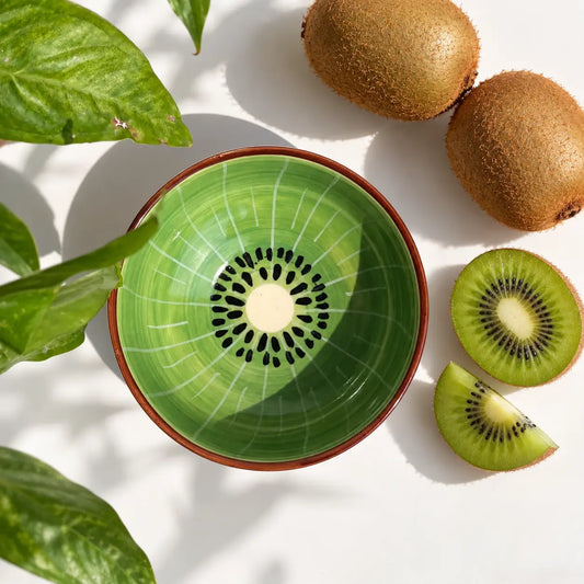 A green ceramic bowl with a kiwi slice pattern inside, styled on a white surface with kiwi fruits and green leaves.