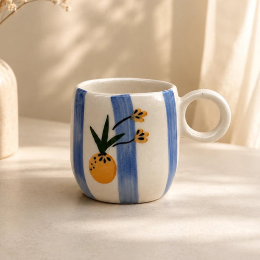 A white ceramic mug with a loop handle, decorated with vertical blue stripes and a painted pineapple motif, set on a table next to a vase of dried flowers in warm natural light.