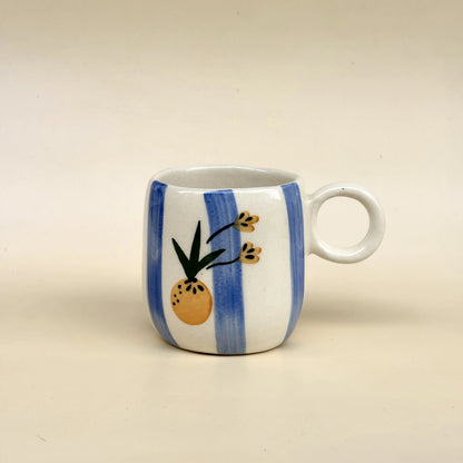 A small white ceramic mug with blue vertical stripes and a hand-painted pineapple design on the front, set against a plain beige background.
