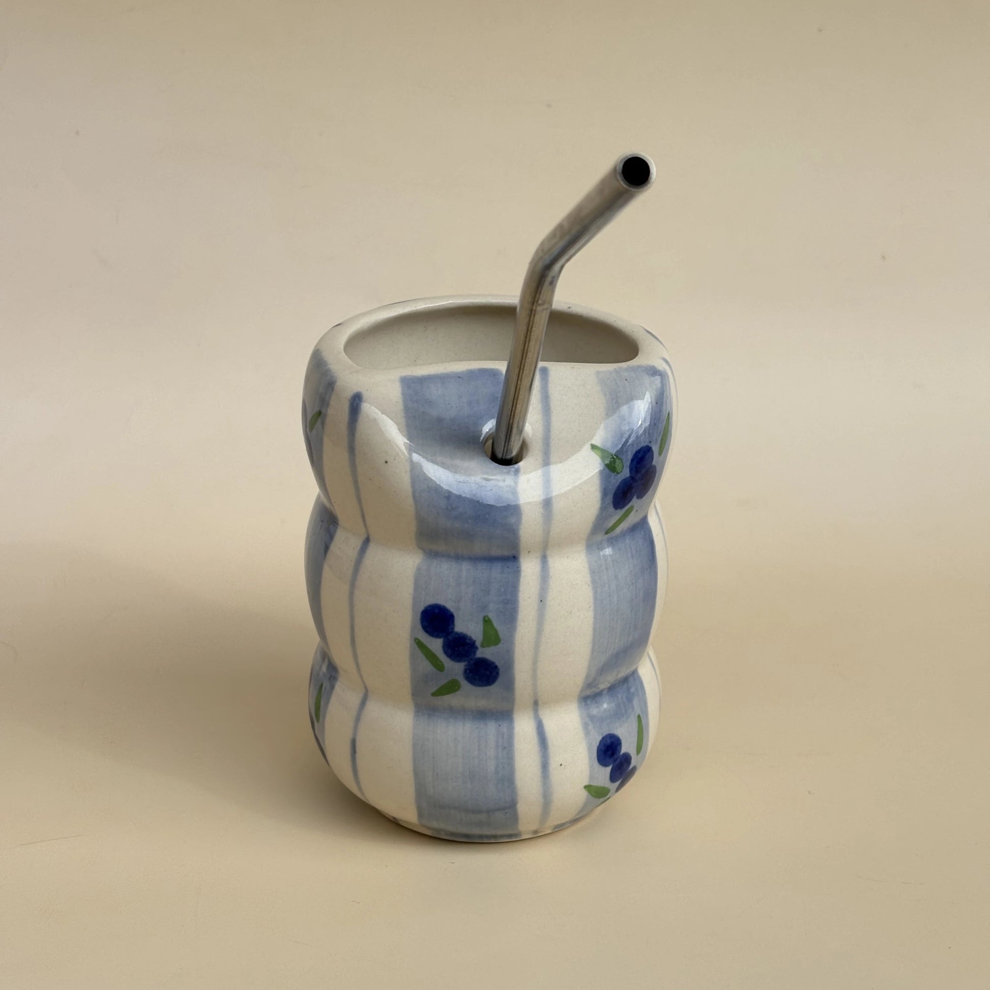 A small ceramic mate cup with a wavy, stacked design in white and light blue, decorated with blue berry motifs, featuring a bent metal straw.
