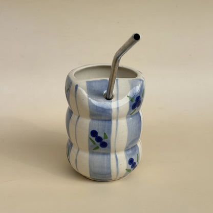 A small ceramic mate cup with a wavy, stacked design in white and light blue, decorated with blue berry motifs, featuring a bent metal straw.