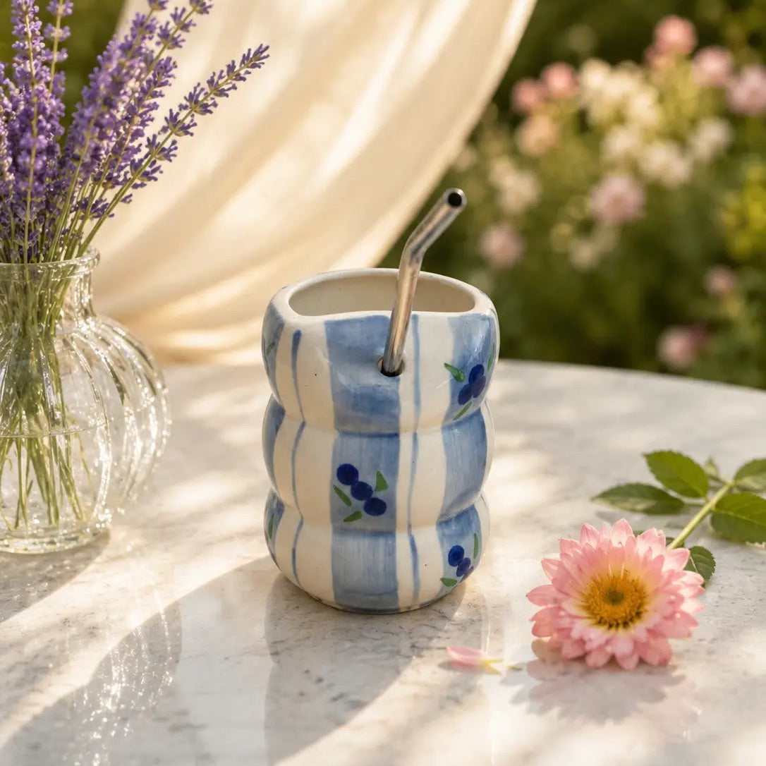 A small ceramic cup with a wavy ribbed design, featuring blue and white stripes and blue berry motifs, with a metal straw. It is styled on a marble table with a pink flower, lavender in a glass vase, and a cream curtain backdrop.