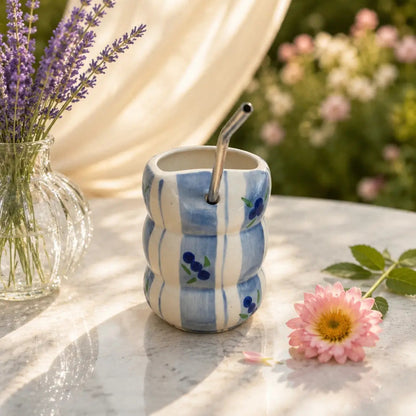 A small ceramic cup with a wavy ribbed design, featuring blue and white stripes and blue berry motifs, with a metal straw. It is styled on a marble table with a pink flower, lavender in a glass vase, and a cream curtain backdrop.