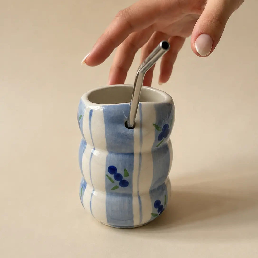 A hand with light pink manicured nails reaches down to adjust a stainless steel straw in a ribbed ceramic cup. The cup features a white and light blue vertical stripe pattern with small blue berry illustrations, set against a solid beige background.