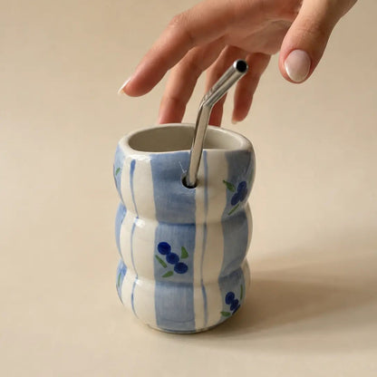 A hand with light pink manicured nails reaches down to adjust a stainless steel straw in a ribbed ceramic cup. The cup features a white and light blue vertical stripe pattern with small blue berry illustrations, set against a solid beige background.
