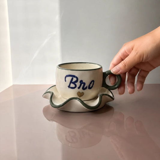 A ceramic coffee cup and saucer set with a hand holding the cup. The cup is cream-colored with a dark green handle and rim, featuring the word 'Bro' in blue script and a small gold heart.