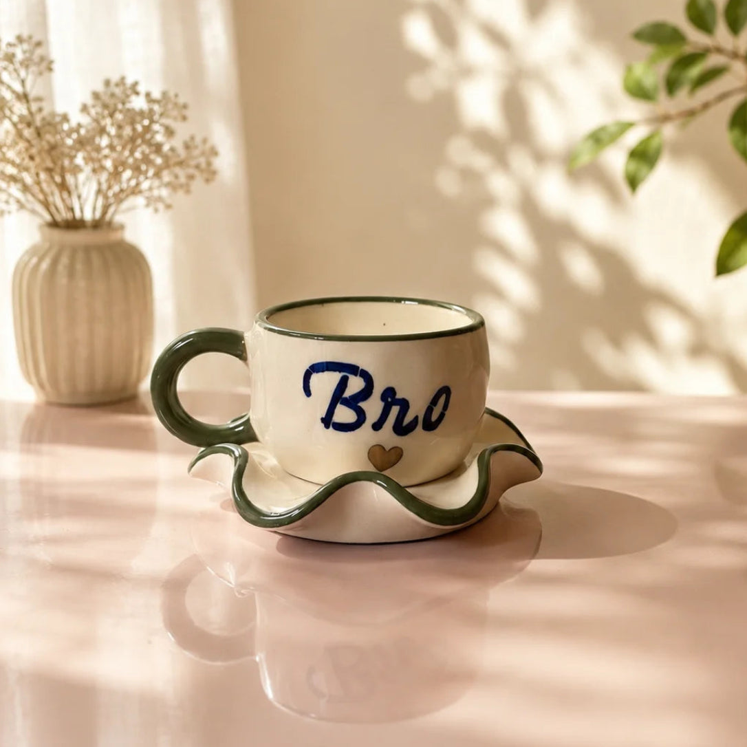 A ceramic coffee cup and saucer set on a pink table, featuring a cream-colored body, dark green wavy rim and handle, and the word 'Bro' written in blue script with a small gold heart. The background shows warm sunlight, leaf shadows, a vase of dried flowers, and a green plant.