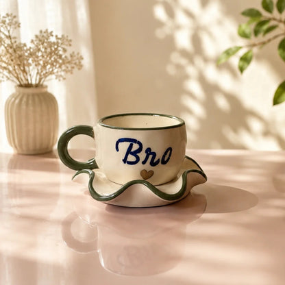 A ceramic coffee cup and saucer set on a pink table, featuring a cream-colored body, dark green wavy rim and handle, and the word 'Bro' written in blue script with a small gold heart. The background shows warm sunlight, leaf shadows, a vase of dried flowers, and a green plant.