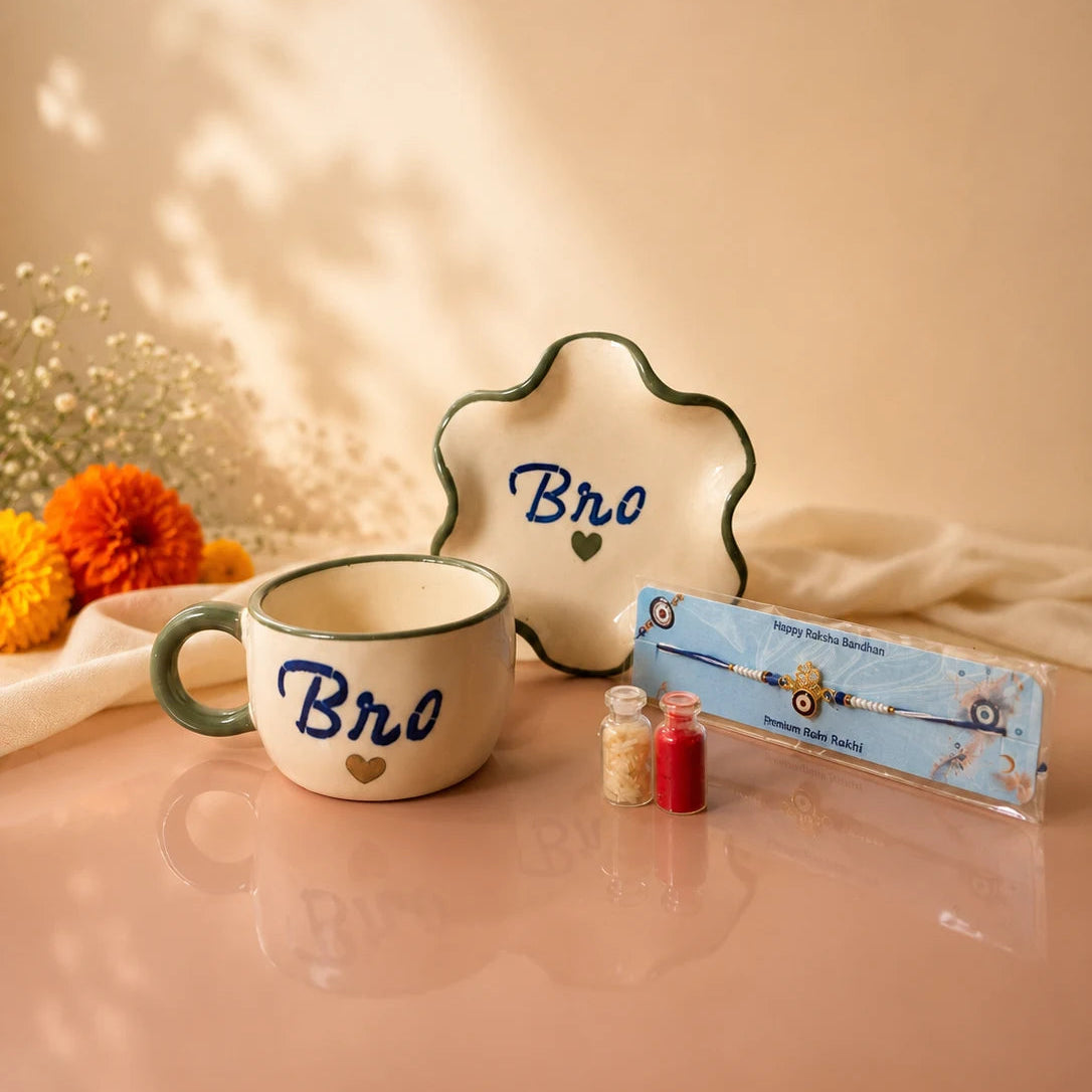 A ceramic mug and matching scalloped plate with the word 'Bro' in blue script and a small heart, alongside a packaged beaded bracelet on a pink tabletop against a warm beige background.