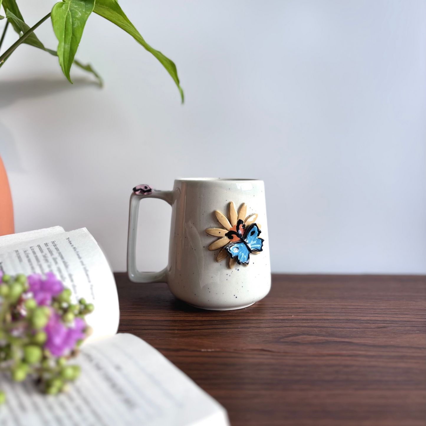 White ceramic mug with decorative elements on a wooden surface next to an open book and plant.