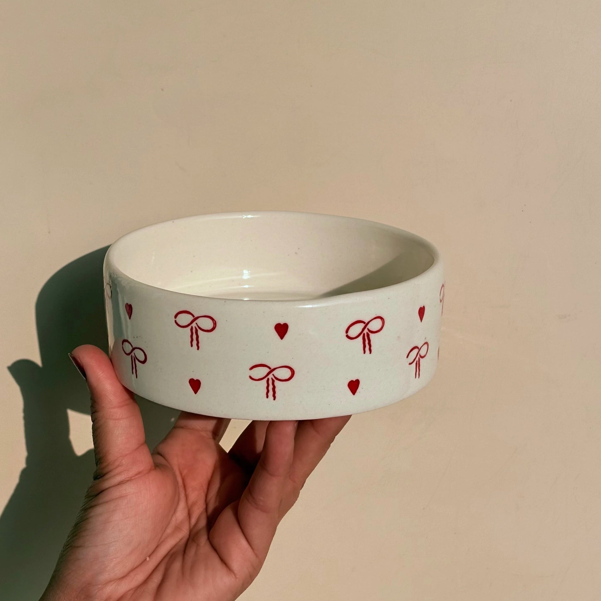White bowl with red heart and bow patterns held by a hand against a beige background