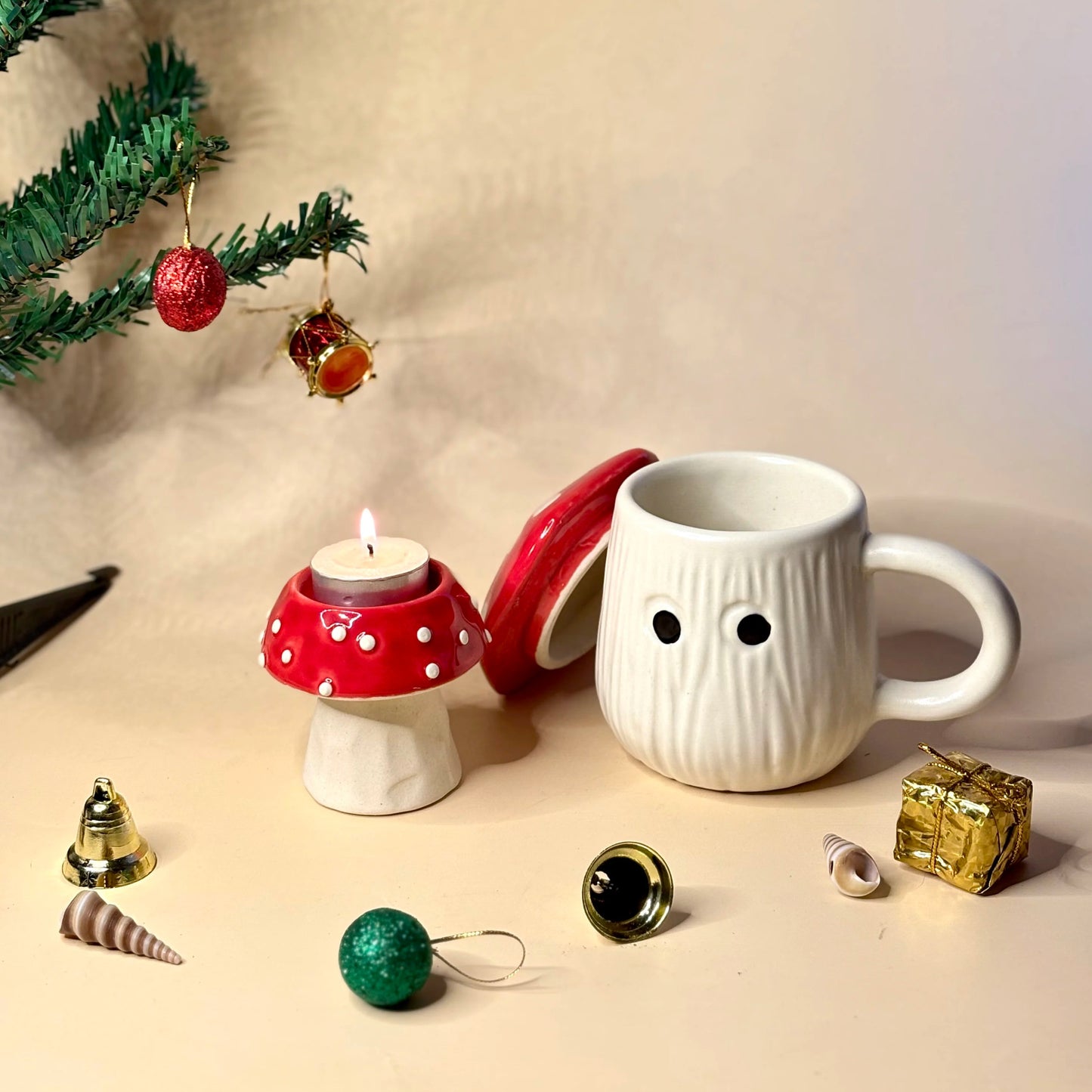 White mug with a face design, red mushroom candle holder, and Christmas ornaments on a beige surface.