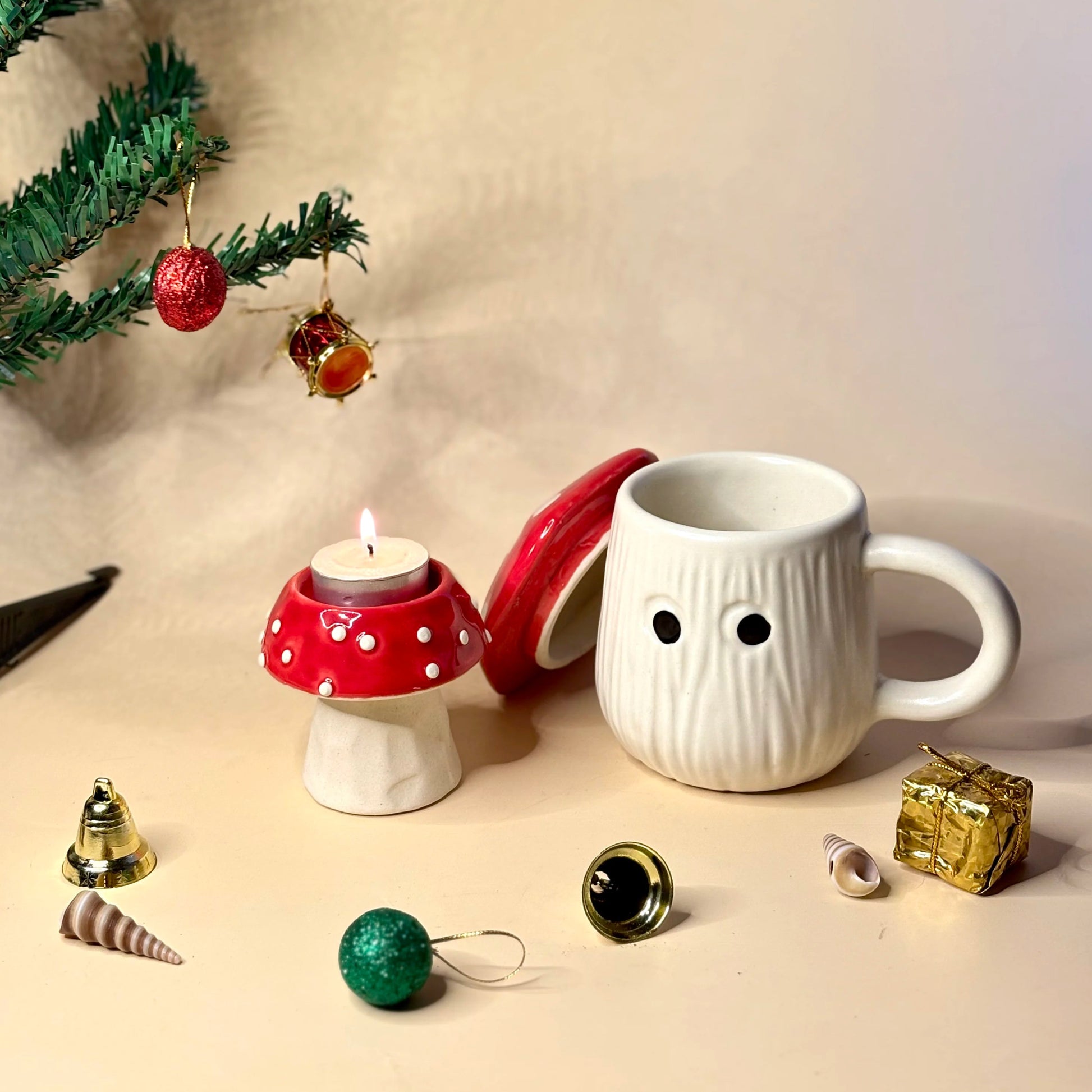White mug with a face design, red mushroom candle holder, and Christmas ornaments on a beige surface.