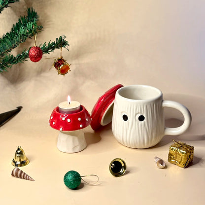 White mug with a face design, red mushroom candle holder, and Christmas ornaments on a beige surface.