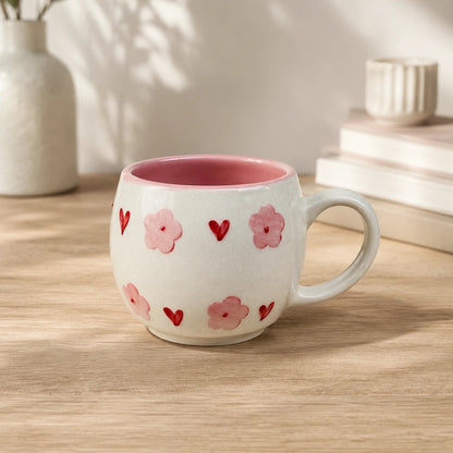 A white ceramic mug with a pink interior, decorated with pink flowers and red hearts, sitting on a wooden table in a softly lit room.
