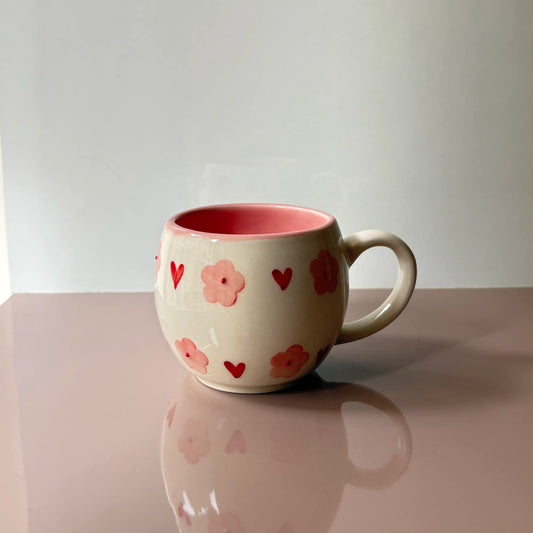 A ceramic mug with a cream-colored exterior decorated with pink flowers and red hearts, featuring a contrasting pink interior and a white handle, set on a reflective pink surface against a neutral light gray background.