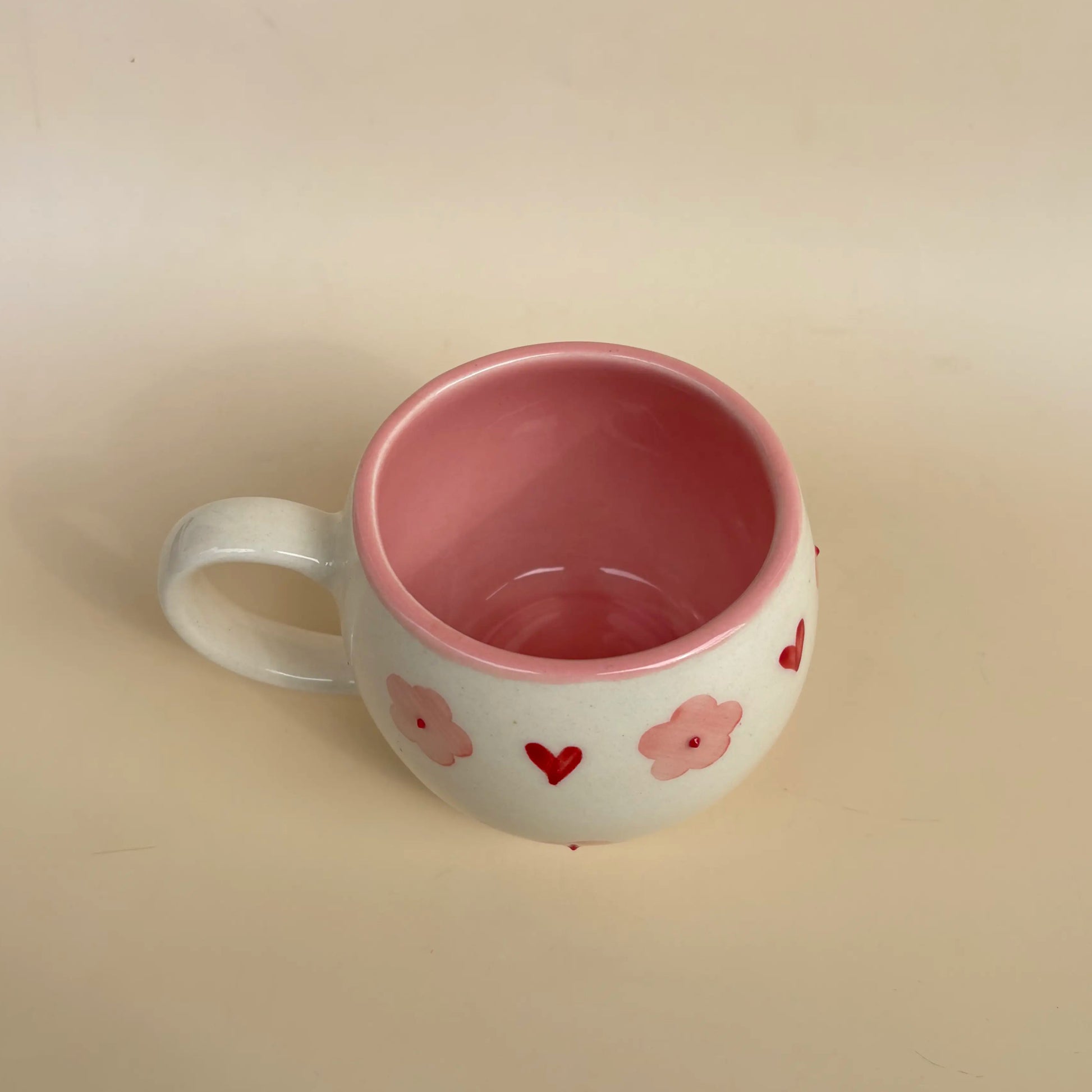 A white ceramic mug with a pink interior and pink heart and flower patterns on the exterior, set against a plain beige background.