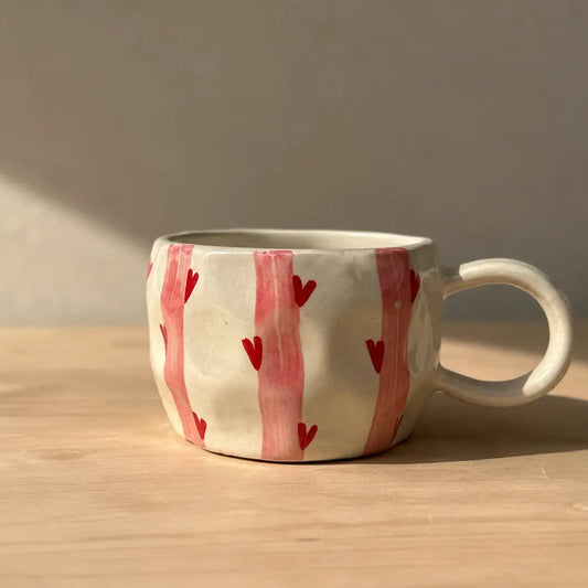 A handmade ceramic mug with a textured off-white glaze, decorated with vertical pink brushstrokes and small red hearts, sitting on a light wooden table under warm natural sunlight.