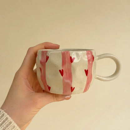 A hand holding a white ceramic mug decorated with vertical pink stripes and small red hearts, set against a plain beige background.