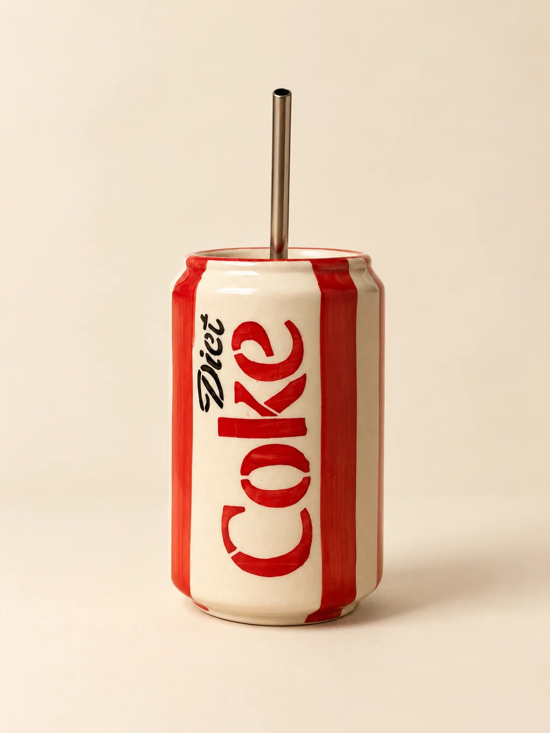 A ceramic tumbler designed to look like a Diet Coke can, featuring red and white vertical stripes and the black 'Diet Coke' logo, with a stainless steel straw inserted at the top, set against a solid beige background.