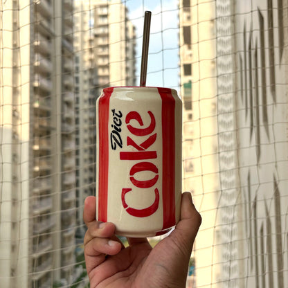 A hand holding a white and red can-shaped tumbler with a metal straw, featuring the 'Diet Coke' logo, with a blurred background of apartment buildings and a net.