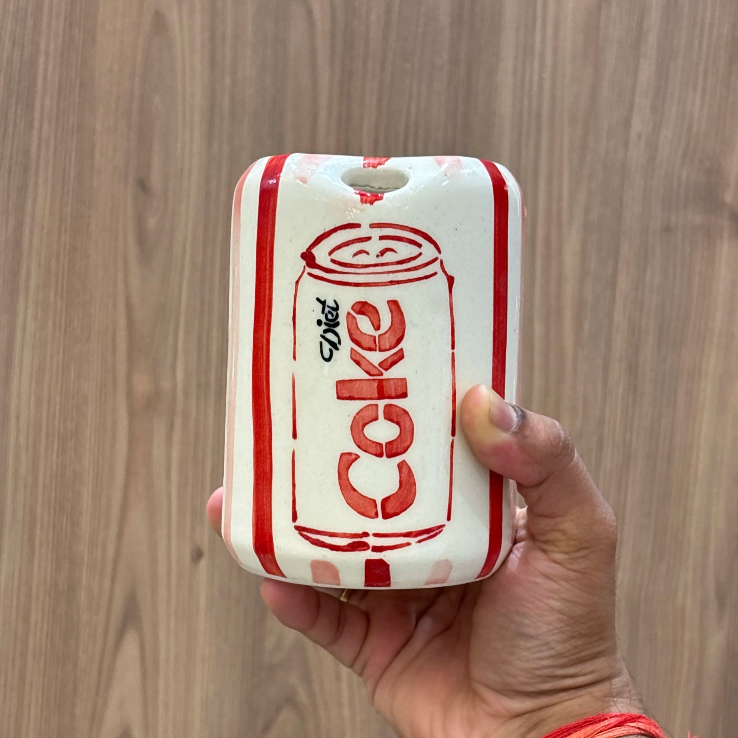 A hand holding a ceramic Diet-Cola can-shaped wall hanging or decor piece against a wood-grain background.