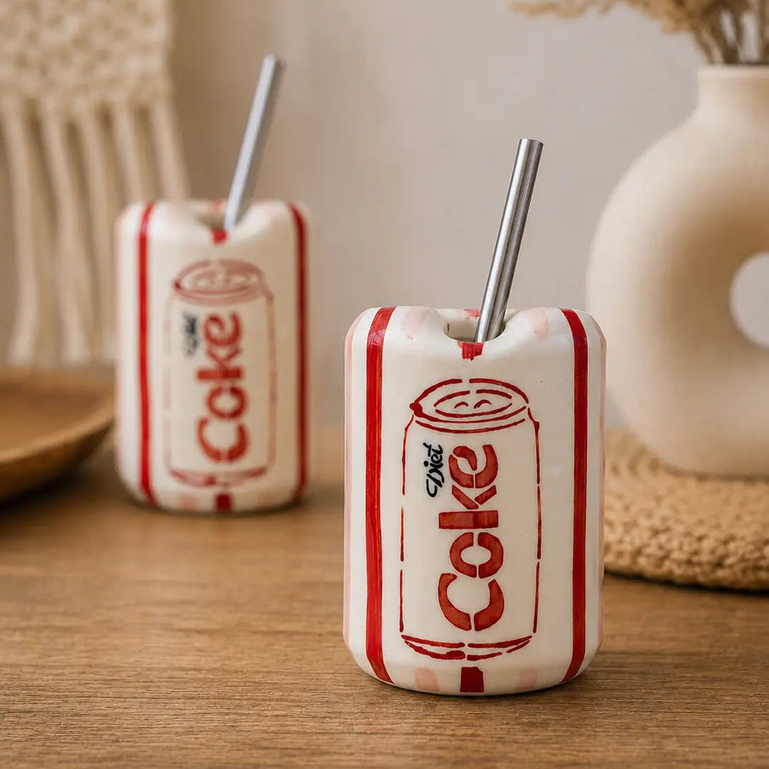 Two ceramic drink holders shaped like vintage Diet Coke cans with red stripes and a metal straw, displayed on a wooden table with a neutral background.