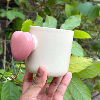 Hand holding a white ceramic cup with a pink handle against a green leafy background