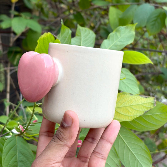 Hand holding a white ceramic cup with a pink handle against a green leafy background