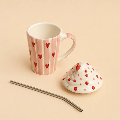 A pink and white striped ceramic mug with red hearts, shown with its matching white lid decorated in red polka dots, alongside a curved stainless steel straw on a solid beige background.