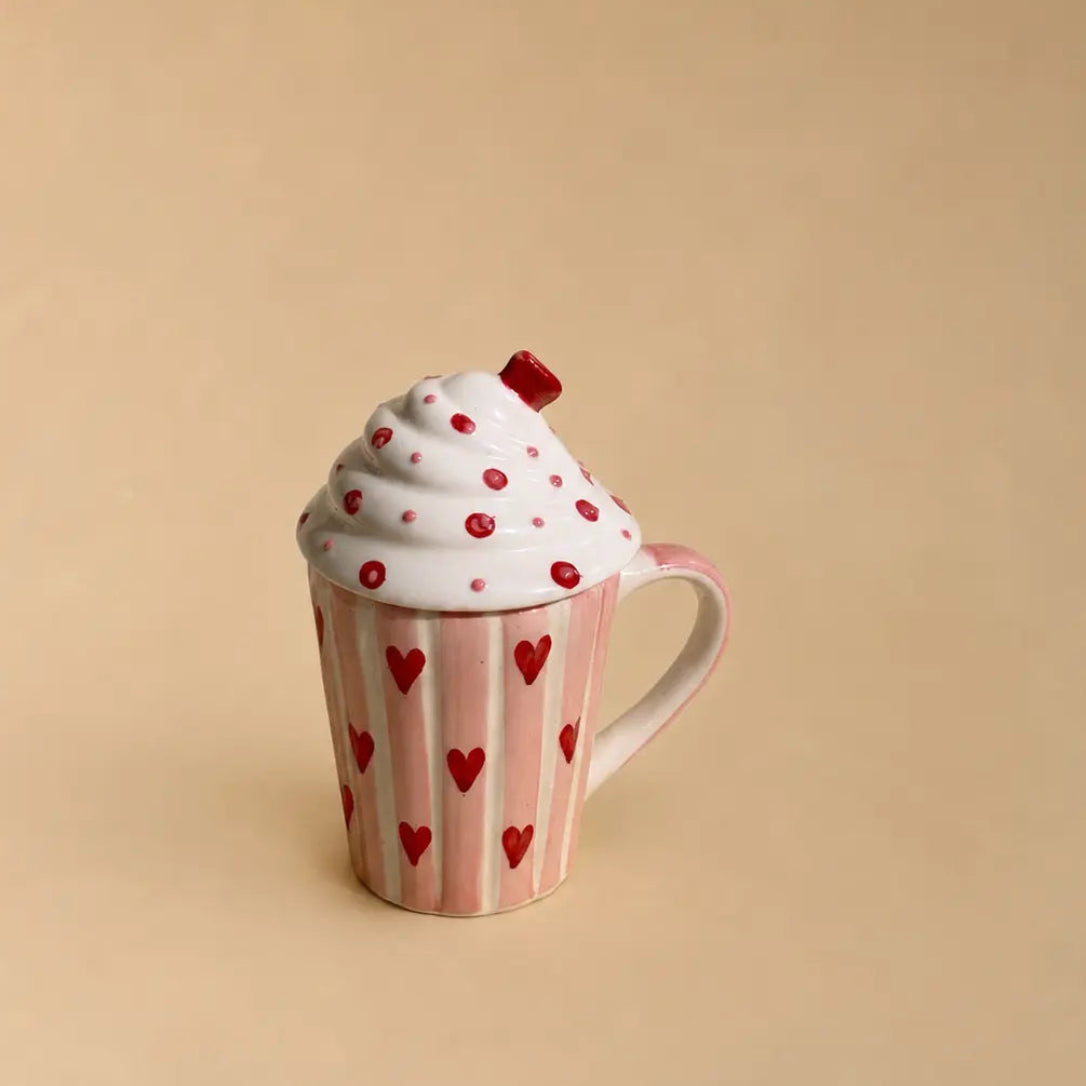 A novelty ceramic mug designed to look like a cupcake, featuring a pink striped wrapper with red hearts, white frosting topped with red hearts and a small red cherry, set against a solid beige background.