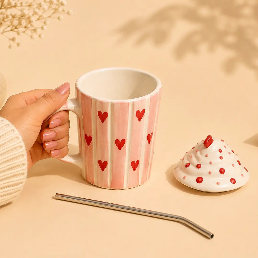 A hand in a cream ribbed sweater holds a pink and white striped ceramic mug decorated with red hearts. The mug is shown on a warm beige table alongside a metal straw, a small white and red speckled cup lid, and dried flowers in a vase.