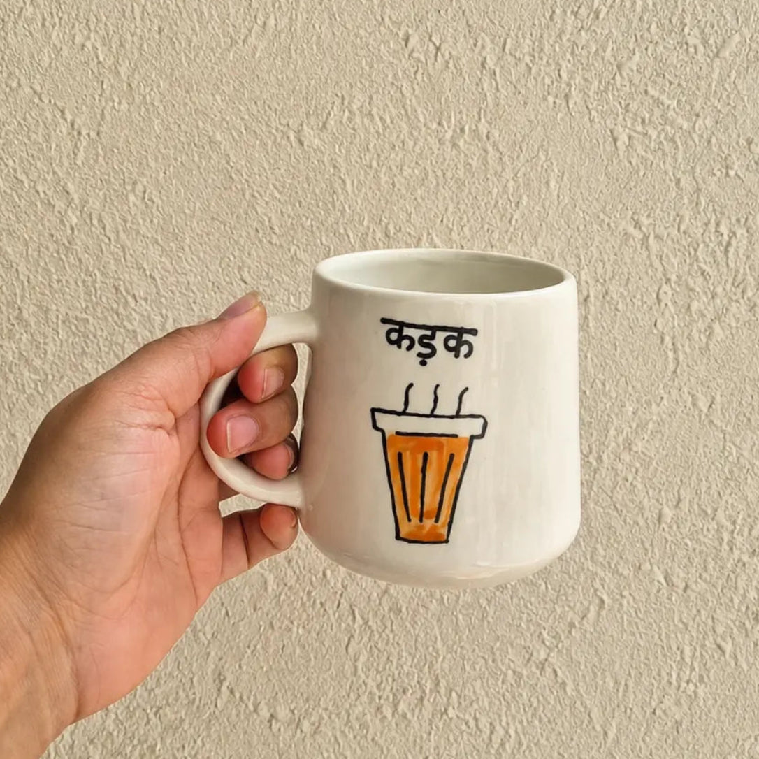 A hand holding a white ceramic mug with a hand-drawn illustration of an orange coffee cup and Devanagari text, set against a textured beige wall.