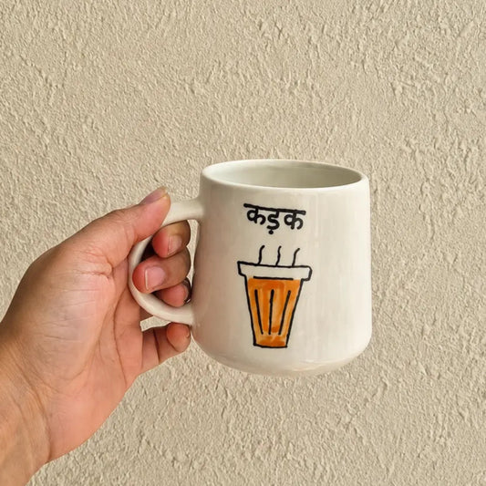 A hand holding a white ceramic mug with a hand-drawn illustration of an orange coffee cup and Devanagari text, set against a textured beige wall.